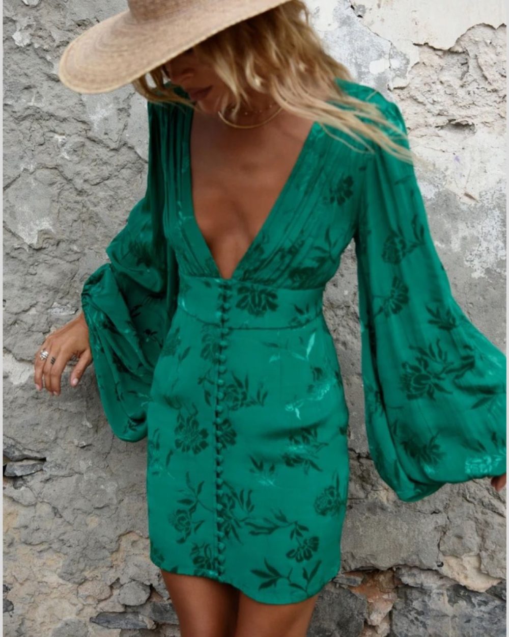 Lady with blonde hair and cream hat in green jacquard mini dress with balloon sleeves against concrete wall looking down.